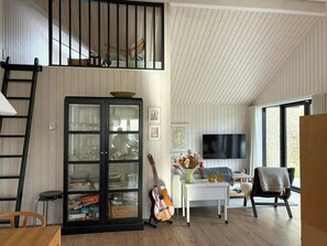 Un espacio habitable tipo loft con una cama en un entrepiso, un mueble con vidrio que contiene vajilla, una guitarra, un televisor y vistas al exterior.