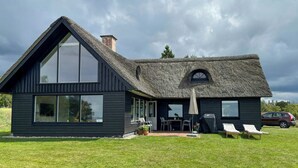 Une maison moderne dotée d’une grande fenêtre, d’un toit de chaume et d’une terrasse avec des meubles d’extérieur.