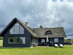 Ein modernes Haus mit einem großen Fenster, einem Strohdach und einer Terrasse mit Gartenmöbeln.