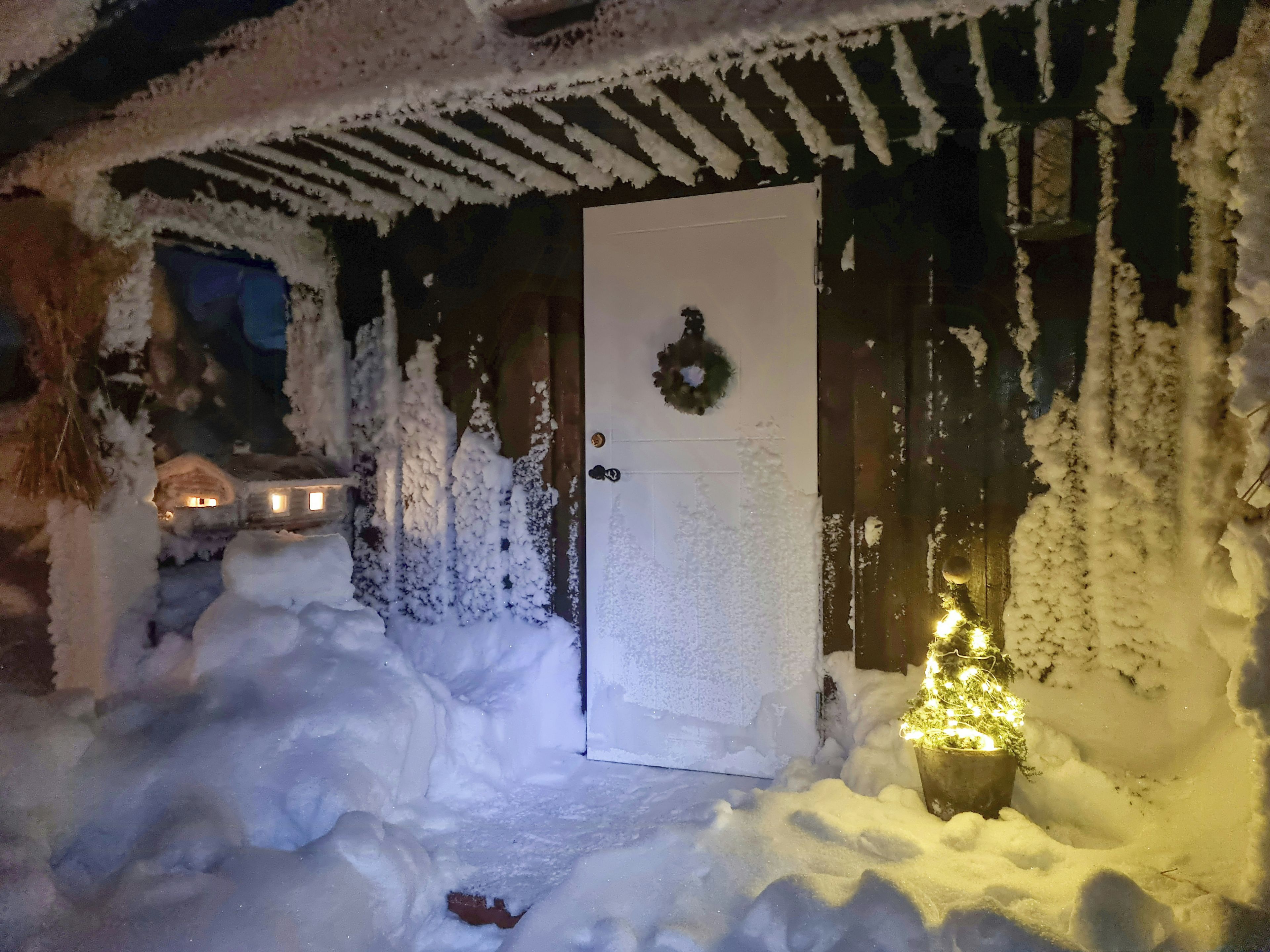 A snow-covered entrance with a door, a wreath, and a lit Christmas tree.