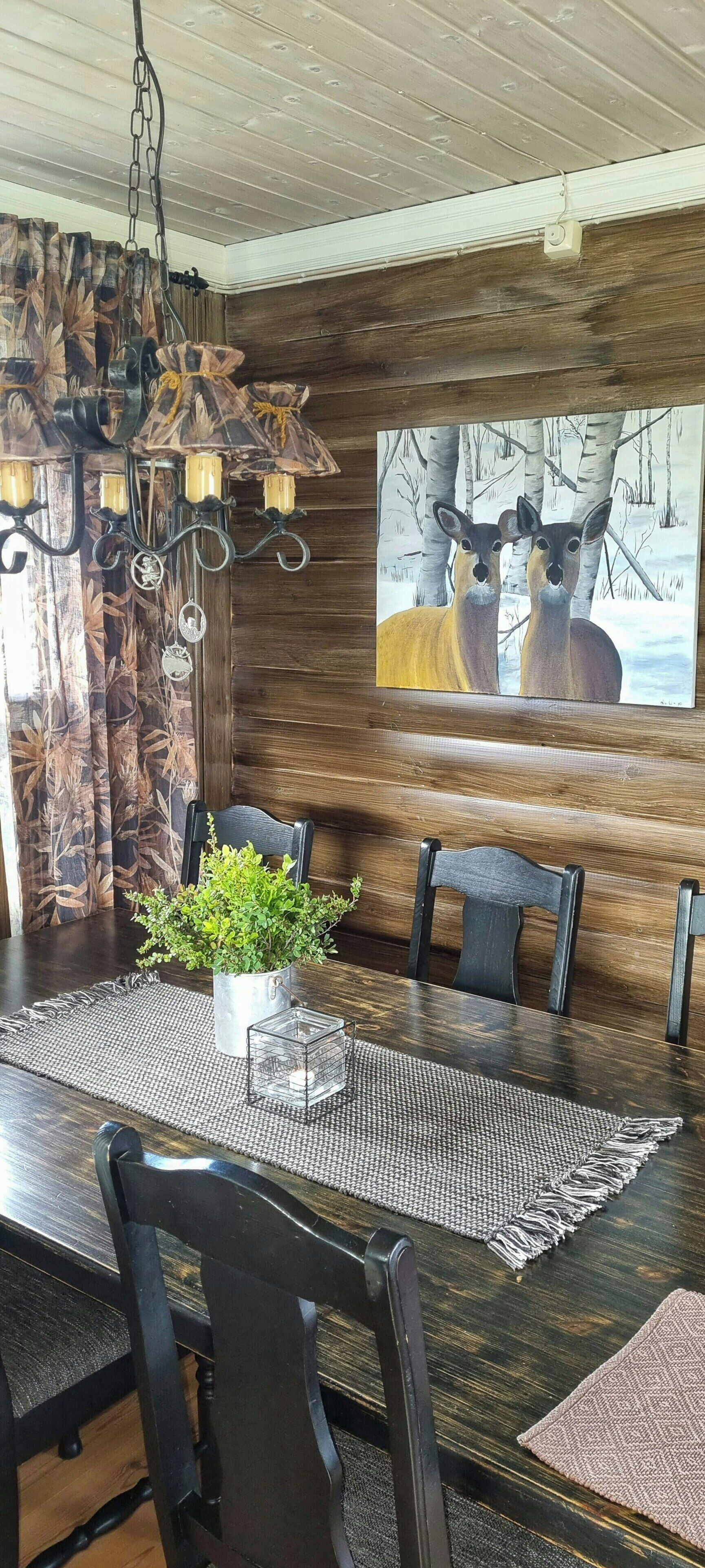 A dining area with a wooden table, chairs, a chandelier, a painting of deer, and a plant centerpiece.