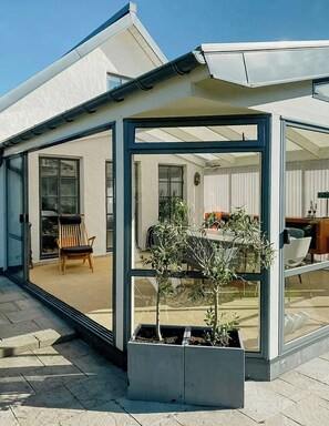 Ein modernes Haus mit einer gläsernen Veranda, einem Holzstuhl und einem kleinen Baumtopf.