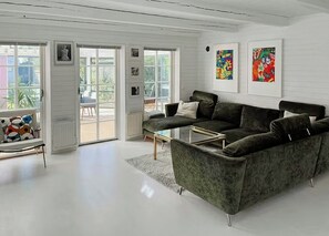 Ein modernes Wohnzimmer mit einem dunkelgrünen Sofa, einem Glastisch und einem weißen Boden. Zwei Türen führen auf einen Balkon, und an den Wänden befinden sich Kunstwerke.