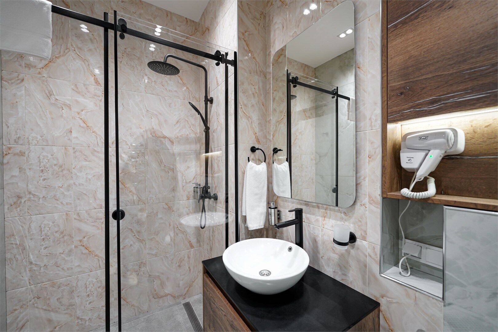 Un baño moderno con ducha de vidrio, lavamanos blanco, espejo y secador de pelo sobre un estante de madera.