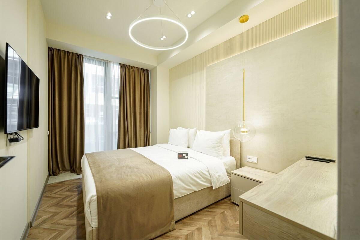 Una habitación de hotel con cama, mesita de noche, televisor y una lámpara moderna.