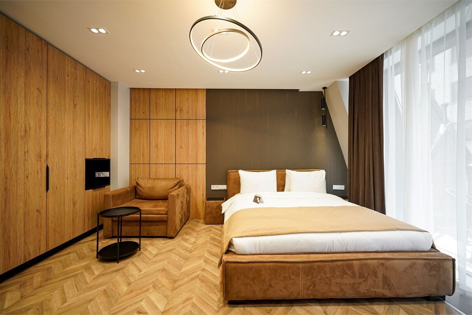 Une chambre d’hôtel moderne dotée d’un grand lit, d’un coin salon avec un fauteuil et une petite table, d’une télévision et d’une grande fenêtre avec des rideaux.