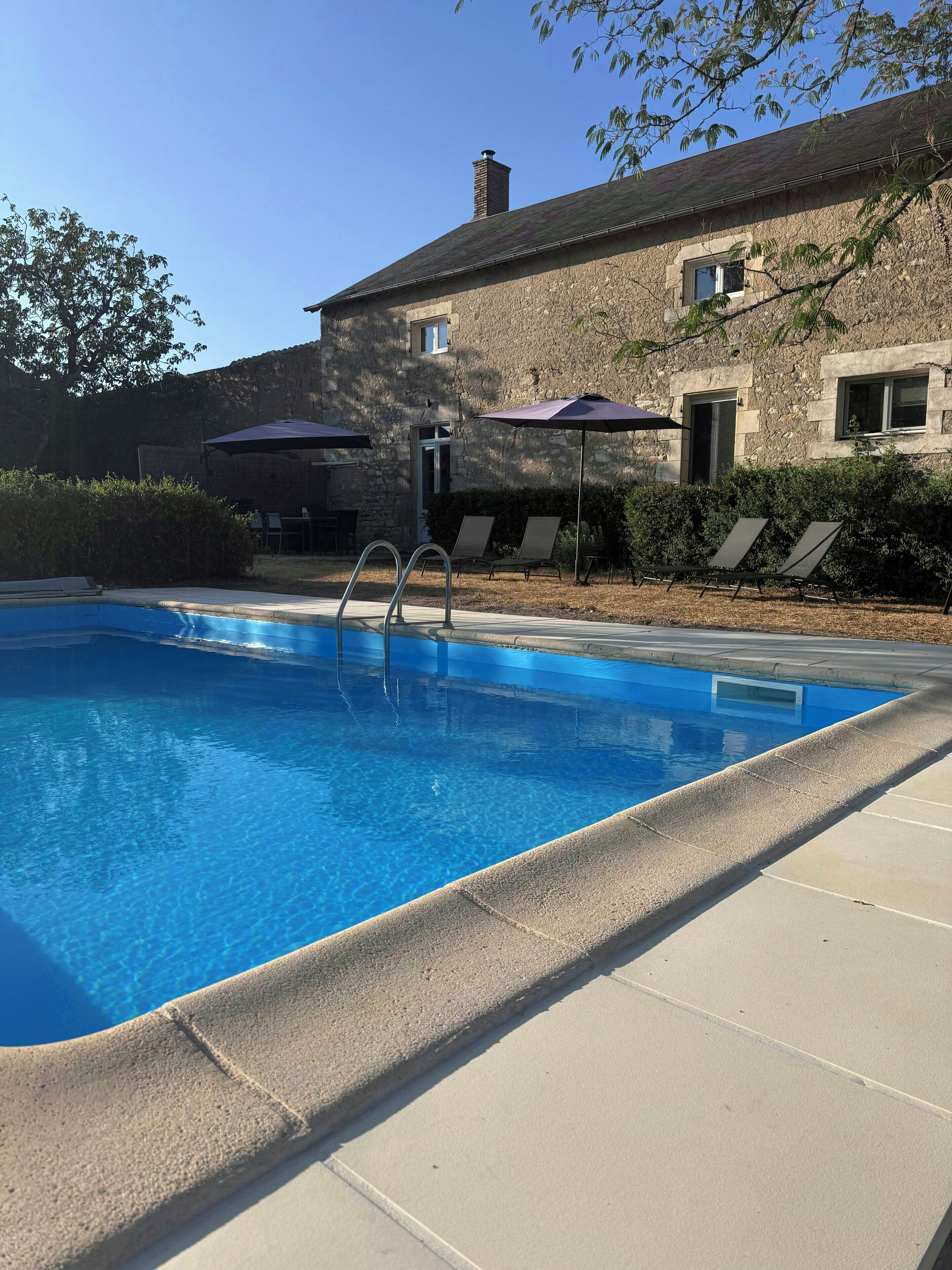 A swimming pool with a stone building in the background, a covered seating area, and a clear sky.