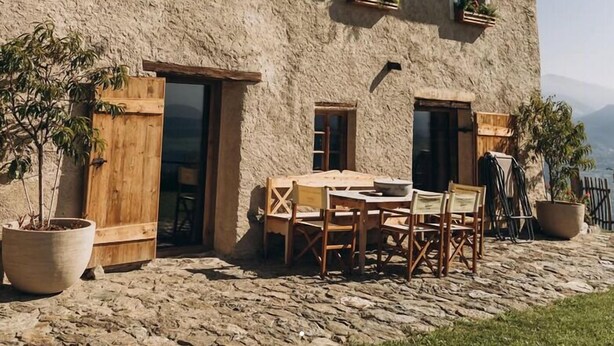 Un edificio rústico de piedra con un balcón de madera, asientos exteriores y macetas con plantas.