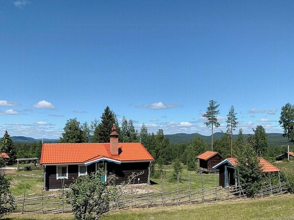 Une cabane rustique avec un toit rouge, entourée d’arbres et de champs ouverts.