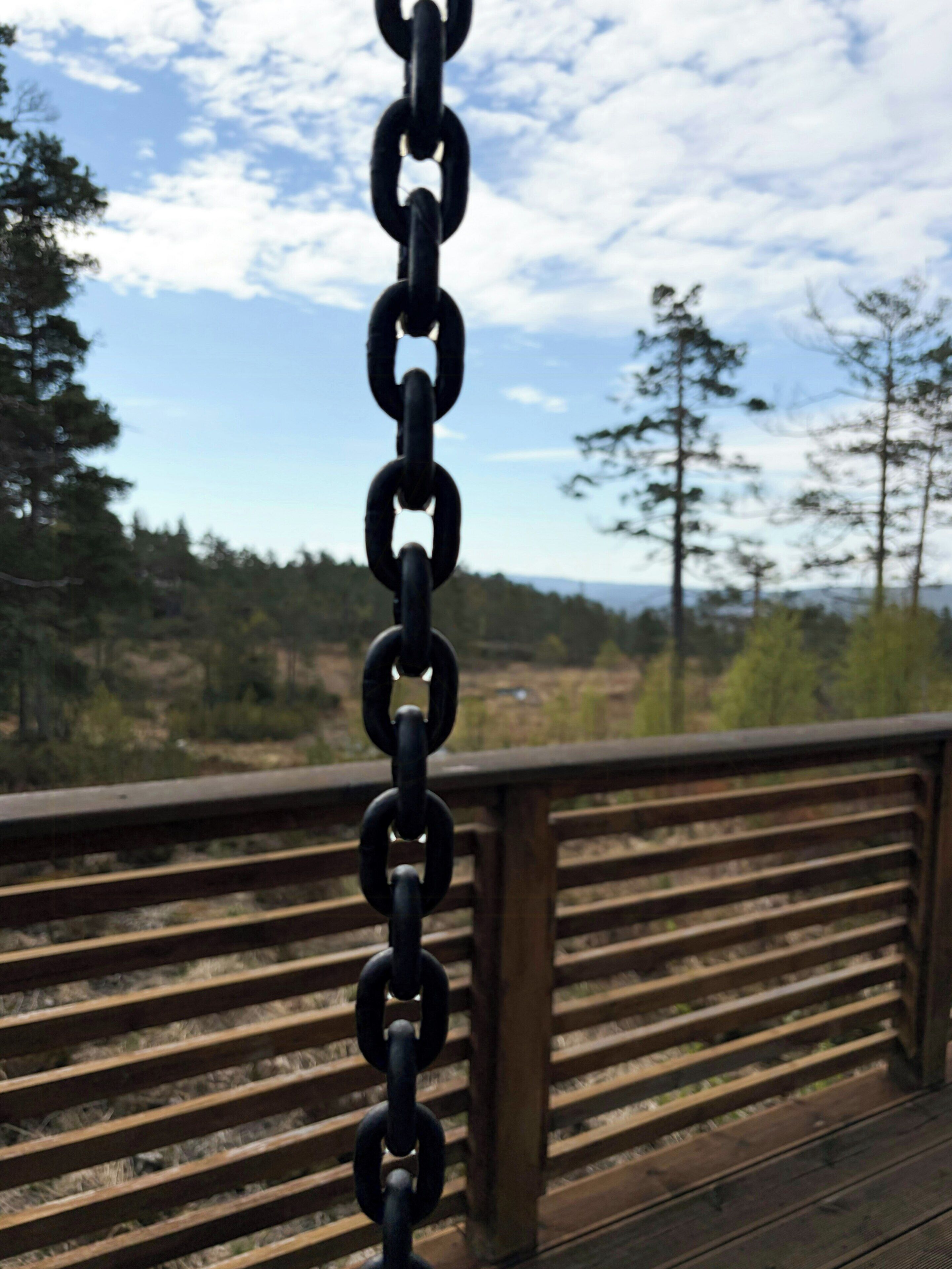 Et hegn af kæder på en træterrasse med udsigt til træer og bakker i baggrunden.