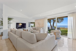 A modern living room with a large beige sectional sofa, a flat-screen TV, and a view of the ocean through large windows.