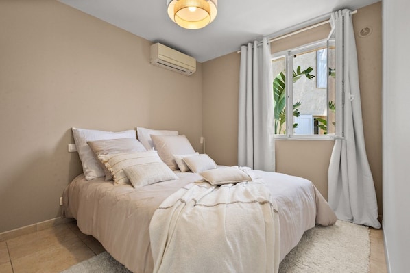 A bedroom with a bed, white bedding, a window with curtains, and an air conditioning unit.