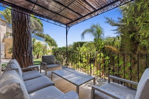 A covered outdoor seating area with a pergola, a table, and chairs, surrounded by a metal fence and lush greenery.