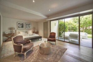 A modern bedroom with a large bed, two armchairs, a coffee table, and a sliding glass door leading to an outdoor patio.