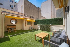 Un patio con césped artificial, una mesa de madera, sillas de mimbre, una pequeña mesa redonda con sillas y un toldo amarillo.