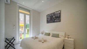 Ein ordentlich bezogenes Bett mit aufgerollten Handtüchern, ein Nachttisch mit Lampe und ein gerahmtes Bild an der Wand.