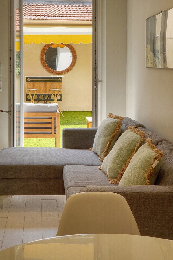 Una sala de estar moderna con un sofá gris, un espejo redondo y vistas a un patio con una mesa y sillas de madera.