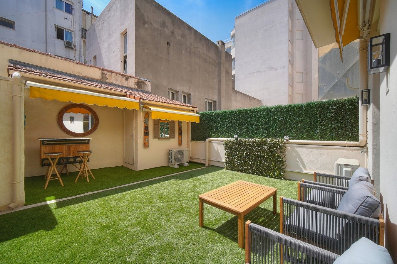 A courtyard with artificial grass, a wooden table, two chairs, a small round table with chairs, and a yellow awning.