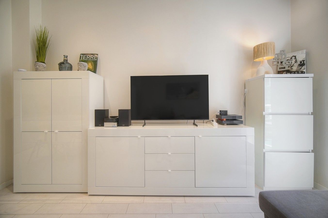 Un salon moderne avec un téléviseur à écran plat posé sur un meuble blanc, un buffet avec des tiroirs et une petite table avec une lampe.