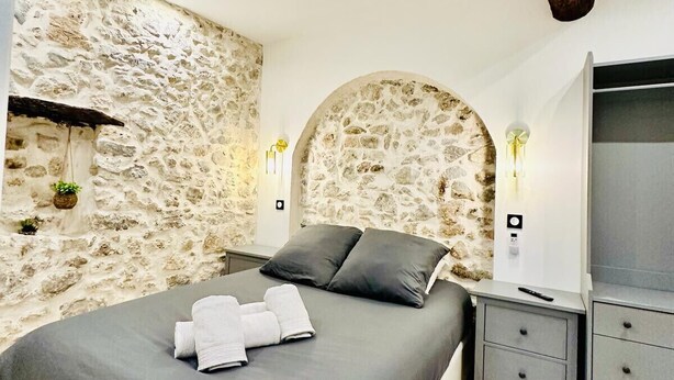 Ein Schlafzimmer mit Steinwand, einem Bett mit grauer Bettwäsche, einem Nachttisch, einem Fernseher und einem Holzbalken.