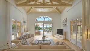 A spacious living room with a high wooden ceiling, a large sofa, a coffee table, a television, and a view of an outdoor patio with a pool. - FRGK Villa Mare Moure Rouge Palm Beach (Cannes)