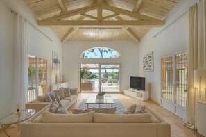 Ein geräumiges Wohnzimmer mit hohen Holzbalken, einem großen Sofa, einem Couchtisch, einem Fernseher und Blick auf eine Außenpatio mit einem Pool.