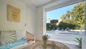 Quarto com sofá, obra de arte na parede, bandeja de vime com saleiro e pimenteiro, e vista para piscina e vegetação lá fora.