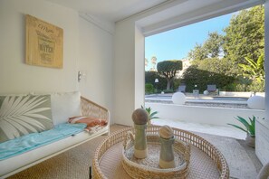 Ein Zimmer mit einer Couch, einem Wandbild, einem Korbtablett mit Salz- und Pfefferstreuern sowie Blick auf einen Pool und Grünflächen.
