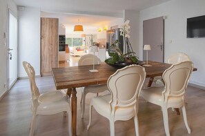 Un comedor con una mesa de madera, sillas blancas, un florero con flores y una cocina al fondo.