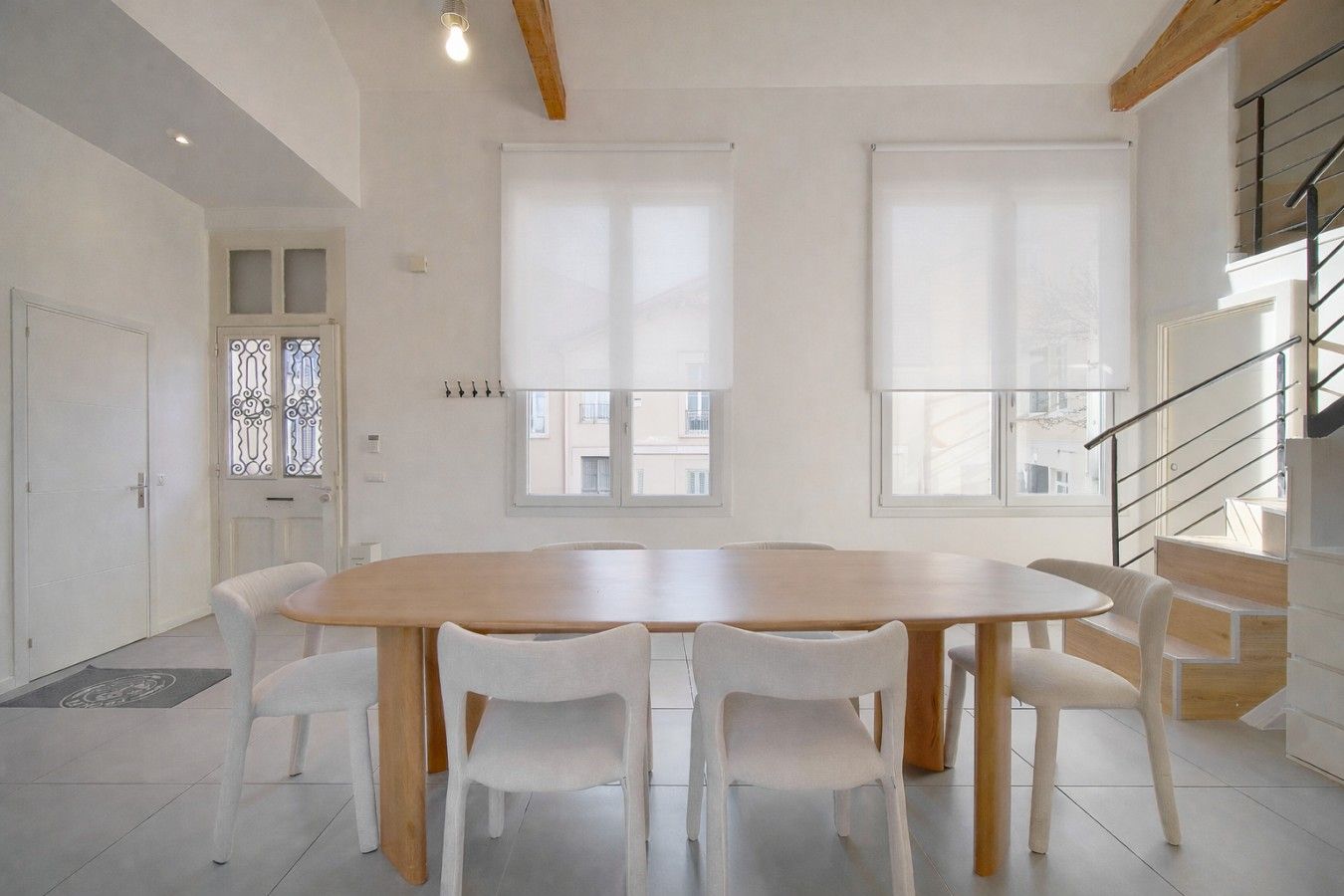 A spacious room with a large wooden table surrounded by chairs, large windows, a staircase, and a door with decorative glass panels.