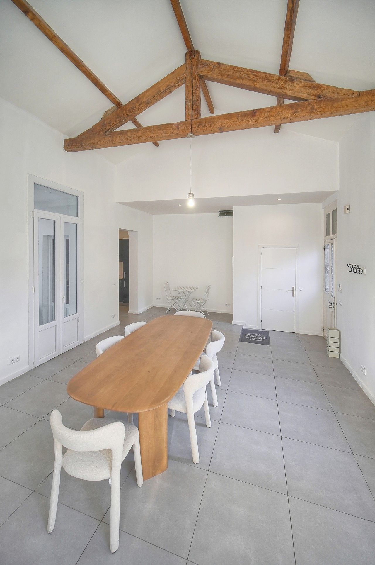 A spacious room with a large wooden table and white chairs, a high ceiling with exposed beams, and a door leading to another room.