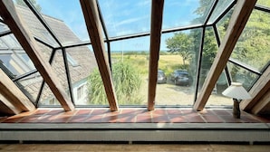 Ein Zimmer mit schrägen Deckenbalken und großen Fenstern, Holzvertäfelung und Blick auf eine grüne Landschaft.