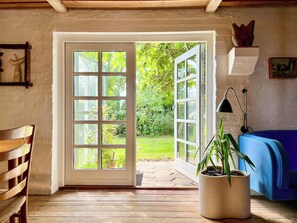 A room with a wooden floor, a white door with glass panels leading outside, a blue sofa, a potted plant, and a lamp. - Artist's house in first row by Smålandshavet (Horslunde)