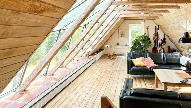 Ein großzügiges Loft mit schrägverdachter Decke, Holzbalken, einem schwarzen Ledersofa, einem hölzernen Schaukelpferd und einer Gitarre im Hintergrund.