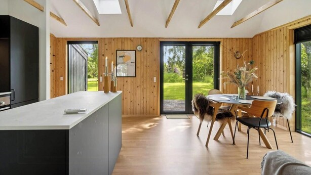 A modern kitchen with a central island, wooden walls, and a large sliding glass door leading to an outdoor area. - Newly renovated holiday home near Torndal beach (Hals)