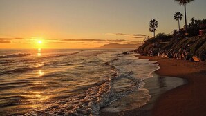 A beach with the sun setting over the ocean, palm trees, and a rocky shoreline. - Villa with rooftop terrace near beach in Marbella (Marbella)