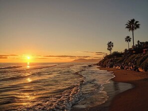 A beach with the sun setting over the ocean, palm trees, and a rocky shoreline. - Villa with rooftop terrace near beach in Marbella (Marbella)