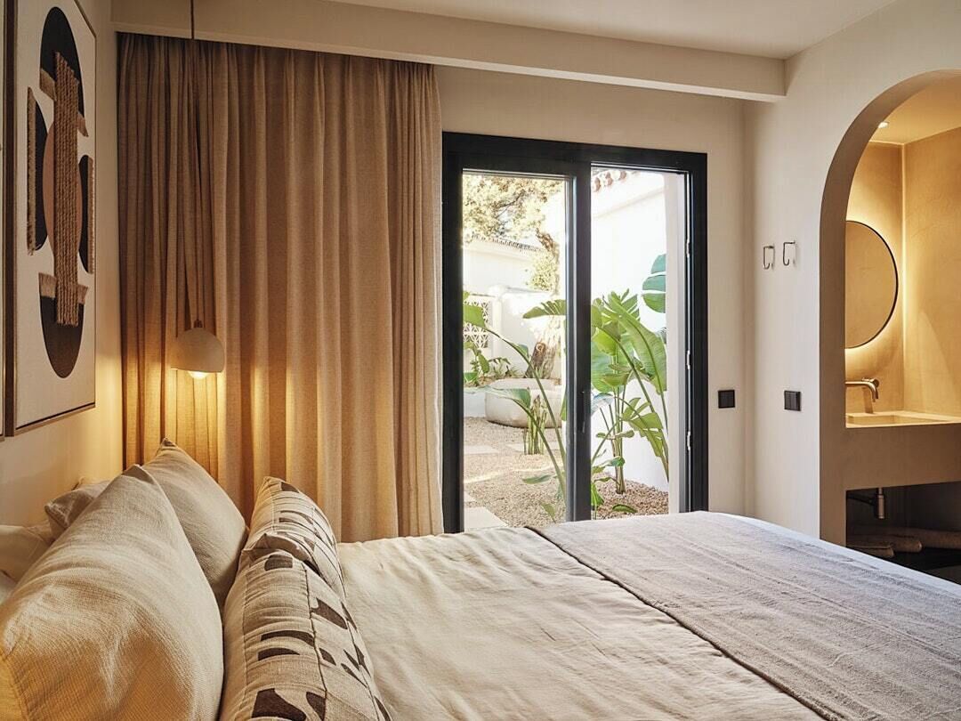 A bedroom with a bed, curtains, a view of the outdoors, and a bathroom with a mirror and sink.