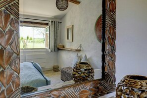 Ein Zimmer mit einem Bett, einem Vorhangfenster, einem Spiegel, einem an der Wand befestigten Regal und einem gewebten Korb.