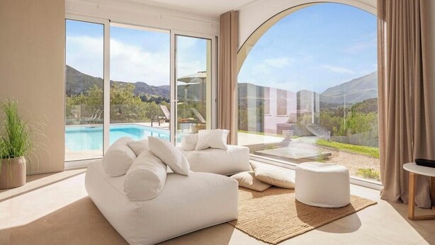 A modern living room with a large window, a white sofa, a small ottoman, and a view of a swimming pool and mountains. - Historic villa overlooking Scopello sea stacks (Castellammare del Golfo)