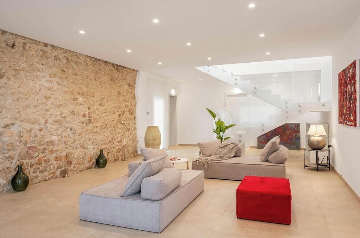 A modern living room with a stone wall, two sofas, a red ottoman, and a staircase leading to an upper level.