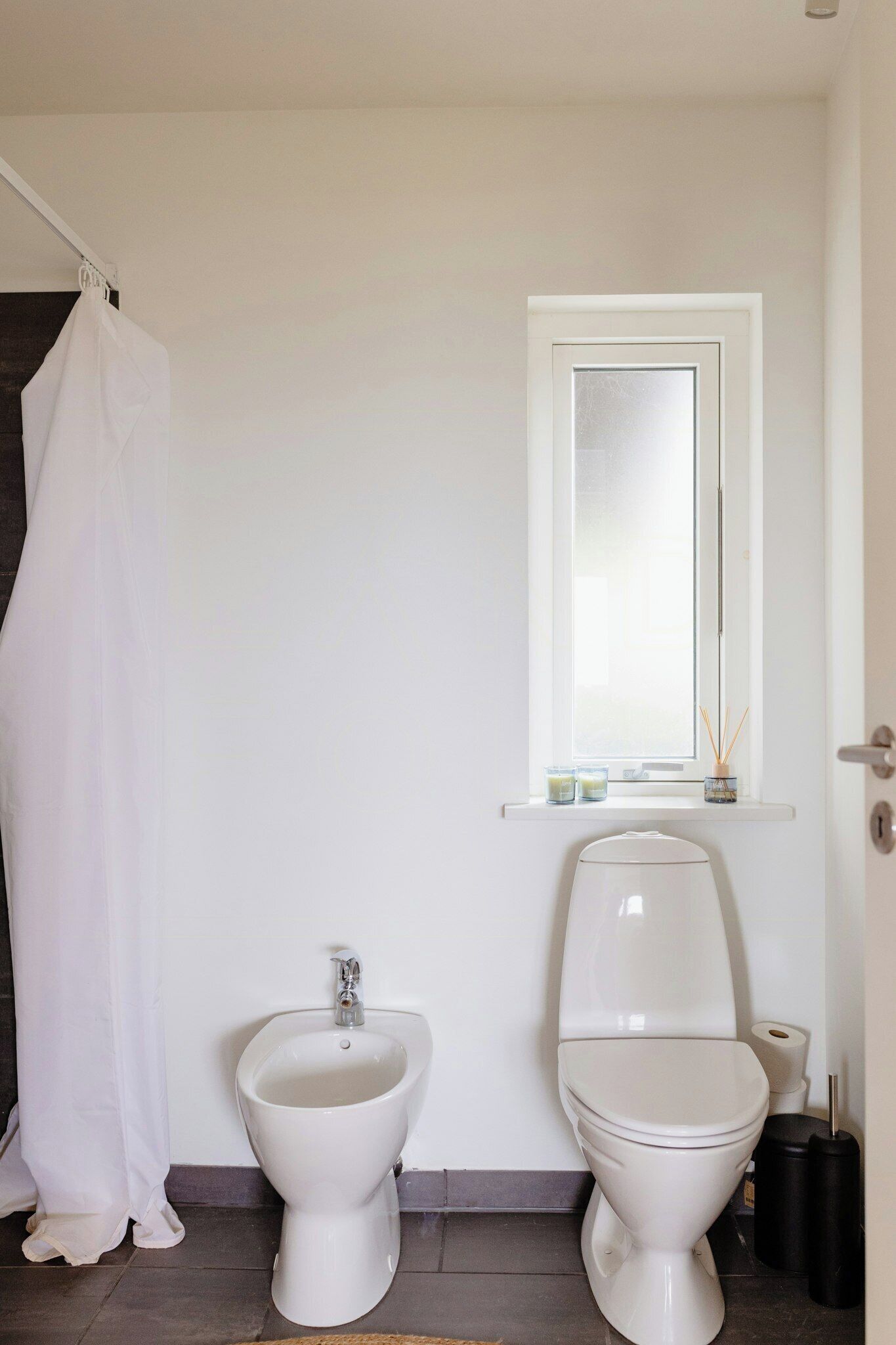 Une salle de bain équipée d’un bidet, de toilettes, d’un rideau de douche blanc et d’une fenêtre avec un miroir.
