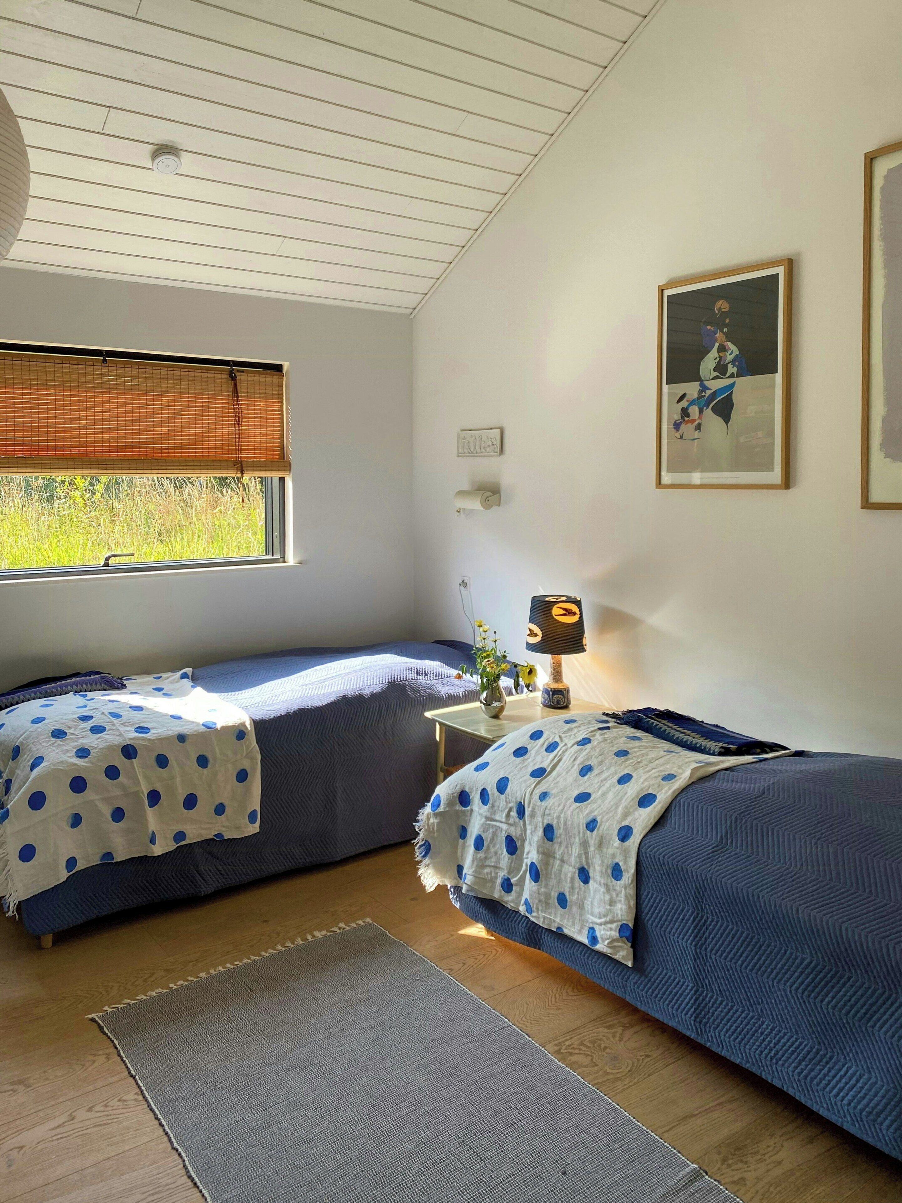 Une chambre avec deux lits, une fenêtre avec des stores, une lampe de chevet, un tableau encadré au mur et un petit tapis sur le sol.