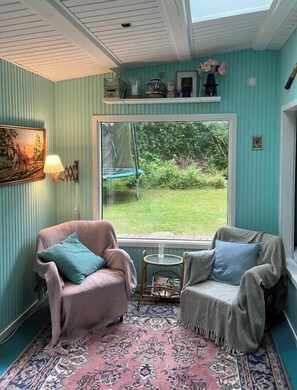 Ein Zimmer mit zwei Sesseln, einem kleinen Tisch, einem gemusterten Teppich, einem Fenster mit Blick auf Grünflächen und einer Wandlampe.