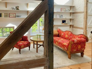 Un salon chaleureux avec un escalier en bois, un canapé à motifs floraux, une table ronde sur laquelle reposent deux verres verts, et une bibliothèque remplie de divers objets.