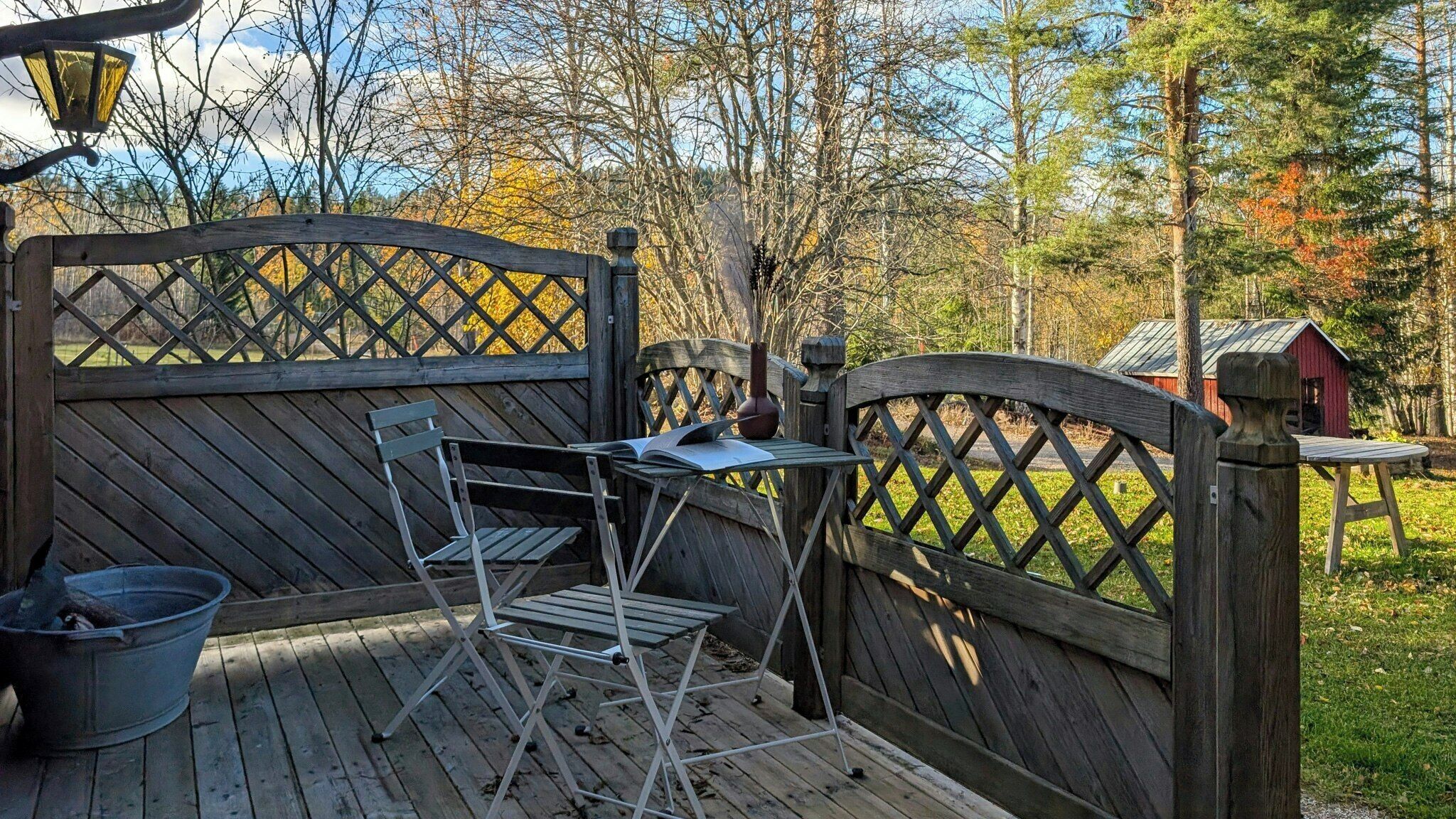 En träveranda med en hopfällbar stol och ett bord i metall, en metallhink och en röd byggnad i bakgrunden.