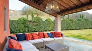 En utomhusplats med sittmöbler, bestående av en soffa och stolar, en pergola ovanför och utsikt över en välskött häck och gräsmatta.
