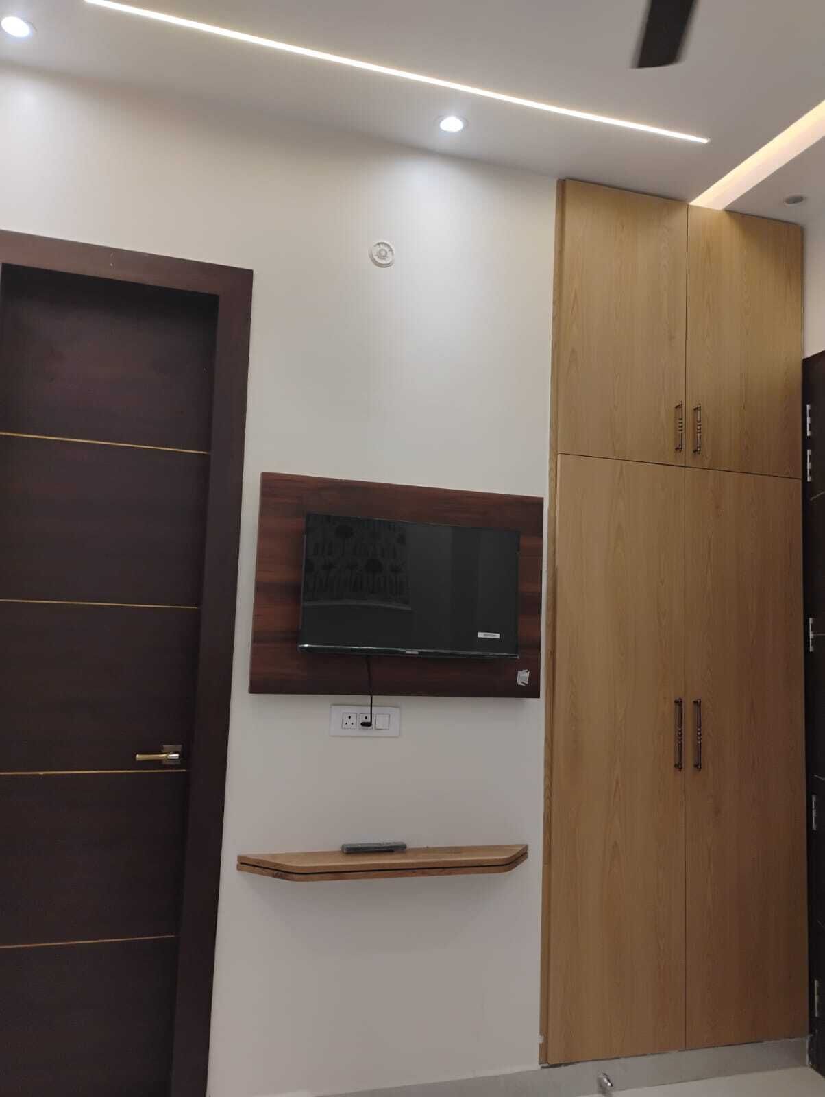 A room with a dark wooden wardrobe, a wooden cabinet, a mounted television, and a small wooden shelf.