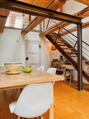 A dining area with a wooden table, white chairs, a bowl, and bananas. A staircase with wooden beams and metal railings is visible in the background. - Modernity in farmhouse with lagoon view (Ummanz)