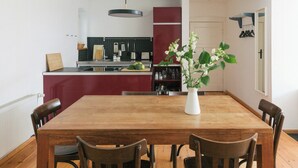 Een eetgedeelte met een houten tafel en stoelen, een vaas met bloemen, en een keuken op de achtergrond.